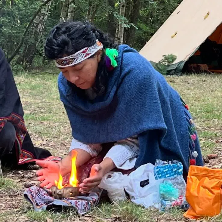 Mar tending a ceremonial fire bowl in the forest with tipi behind